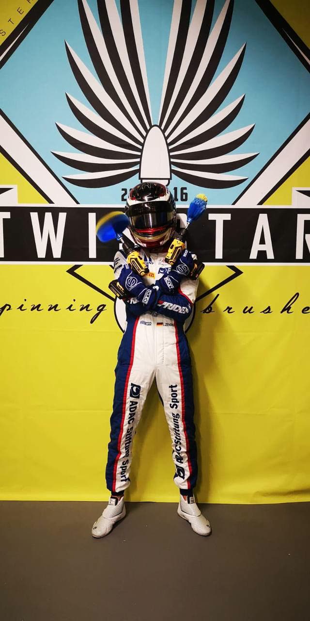 Lirim Zendeli in his race suit in front of the giant TwistStar emblem wall, holding two drill brushes crossed over his chest.